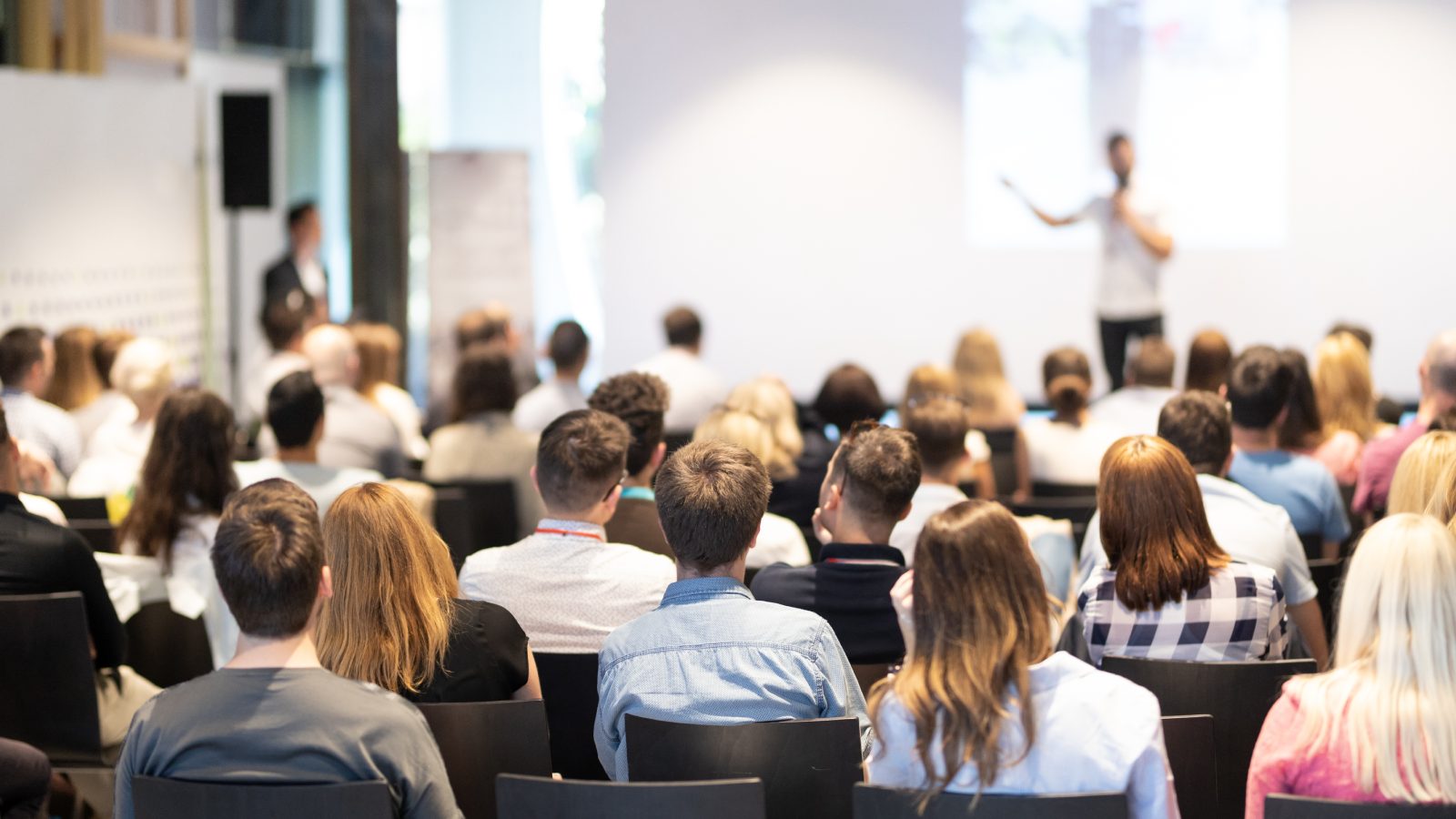 A speaker stands at the front of a large room filled with people, gesturing to the presentation screen behind him.
