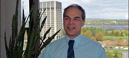 A smiling man in a light blue shirt and dark blue tie sits by a window with a tall office building and scenic park visible in the background. A large potted plant is beside him.