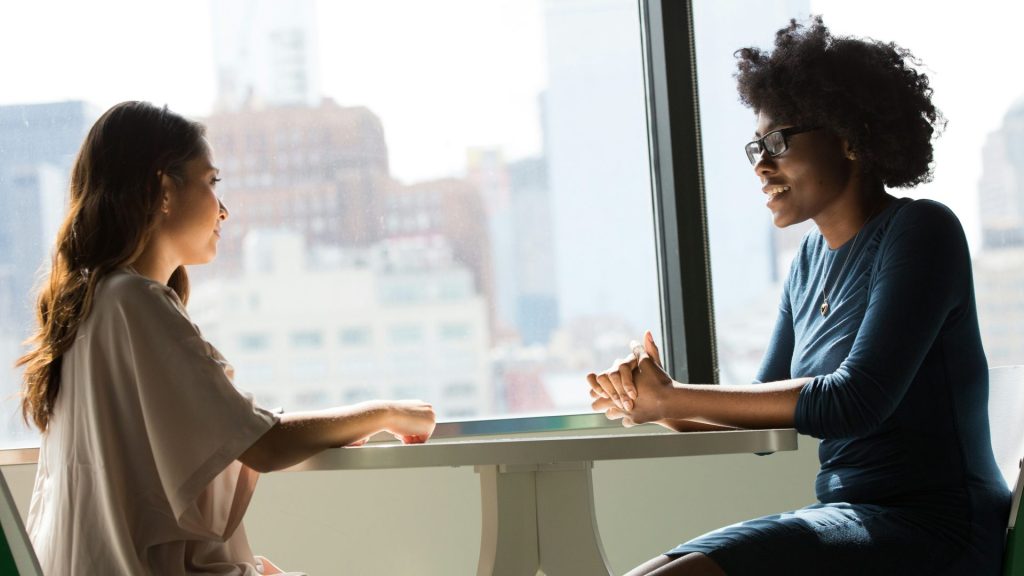 Two professional women in business outfits sit across from one another at a table in front of a window.