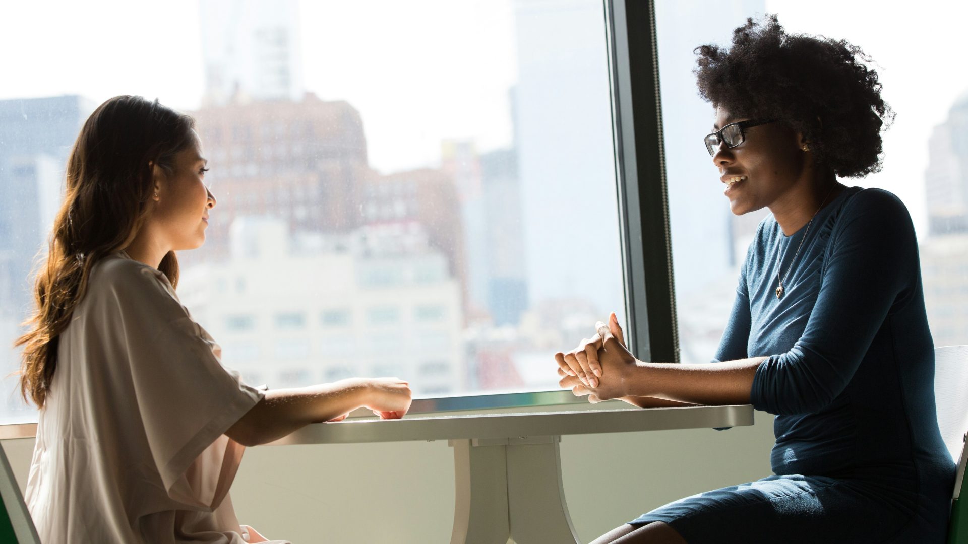 Two professional women in business outfits sit across from one another at a table in front of a window.