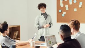 A three-person team sits around a table with their laptops open and their notes spread out in front of them. A fourth-team member stands in front of a bulletin board full of sticky notes and addresses their colleagues.
