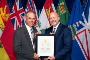 Tony Labillois in a suit smile while holding a framed Career Excellence Award certificate in front of Canadian provincial and territorial flags.